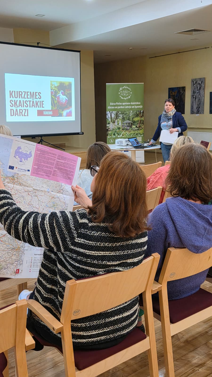 Seminārā dalībnieki klausās prezentāciju par Kurzemes skaistajiem dārziem; priekšplānā redzama karte, bet fonā – Dārza Pērles informatīvais stends.