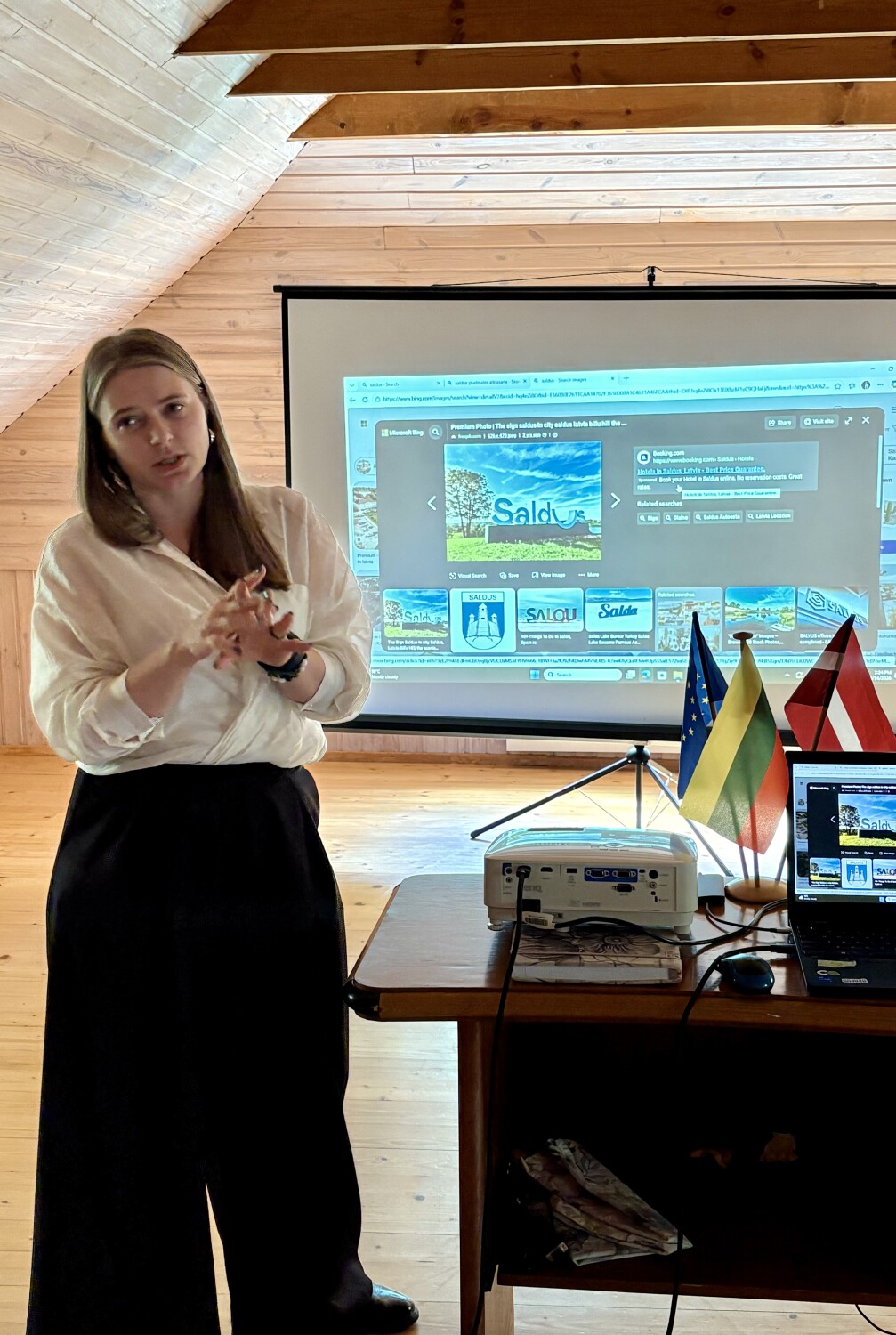 Sieviete koka telpā vada prezentāciju pie projektora ekrāna, uz kura redzams Saldus attēlu meklējums; uz galda novietoti projektors, dators un Latvijas, Lietuvas un ES karodziņi.
