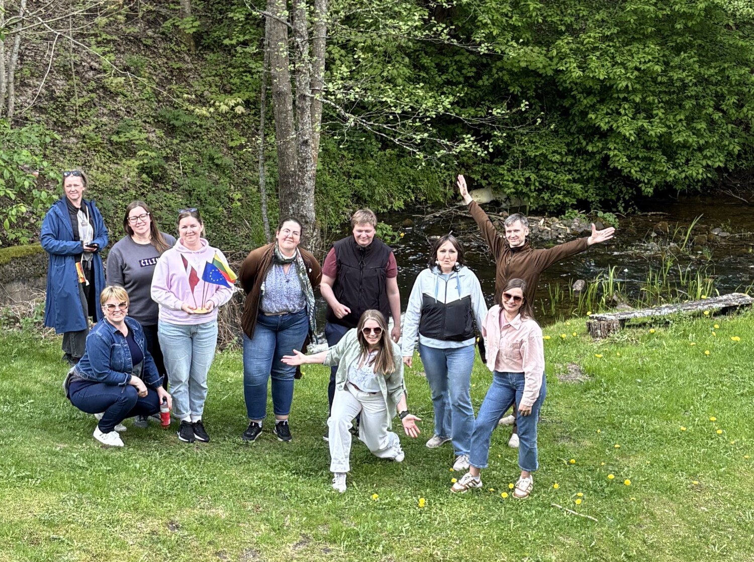 Cilvēku grupa pozē zaļā pļavā pie nelielas upes meža ielokā, smaidot un mājot kamerai; vienai dalībniecei rokās ir Latvijas un Eiropas Savienības karodziņi.