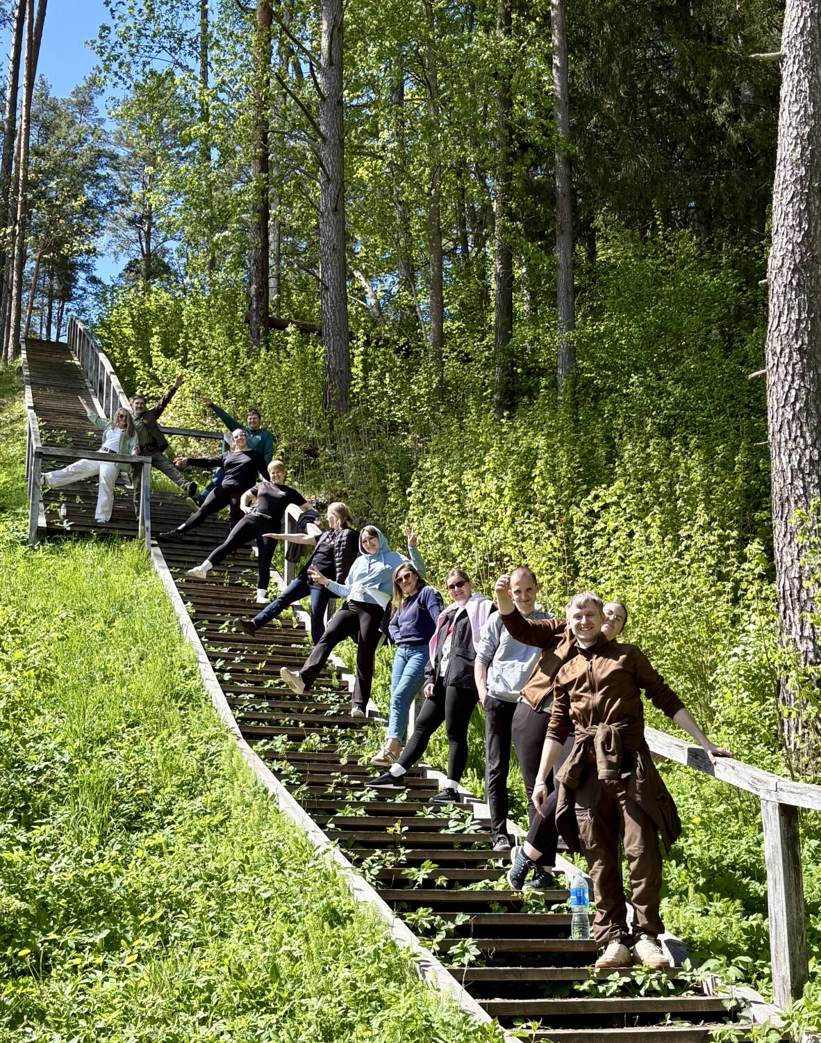 Cilvēku grupa saulainā dienā pozē uz garām koka kāpnēm mežainā nogāzē, smaidot un mājot kamerai, apkārt redzami zaļi koki un pavasarīga daba.