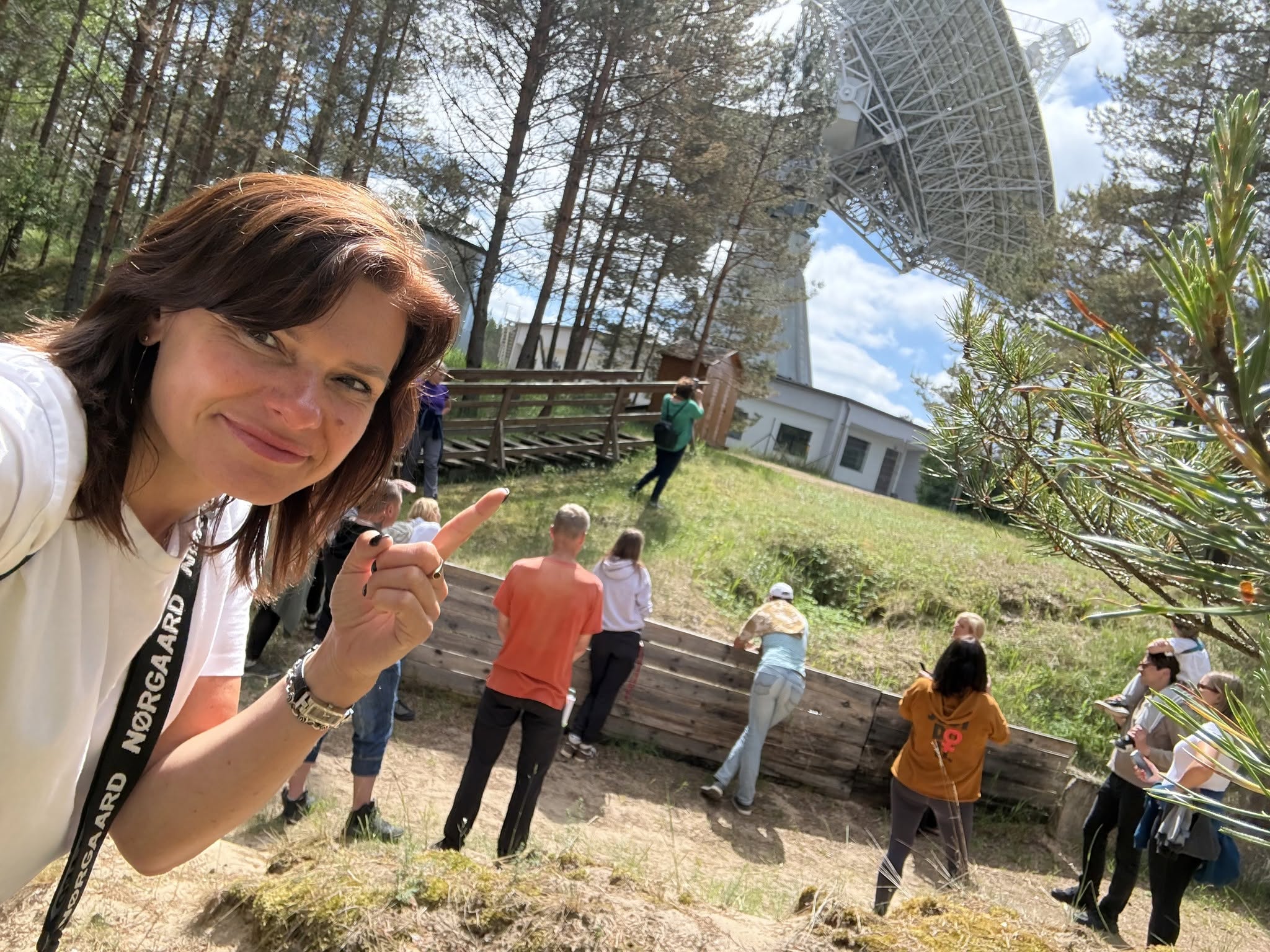 Dalībnieki apmeklē Irbenes radioteleskopu, savukārt priekšplānā sieviete norāda uz iespaidīgo objektu.