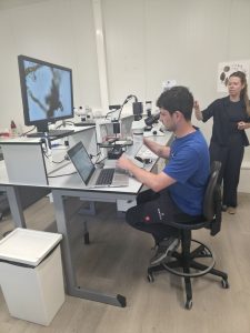 Vīrietis laboratorijā pēta paraugu mikroskopā un vēro attēlu monitorā, kamēr sieviete skaidro izpētes procesu.