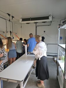 Apmeklētāji apskata laboratorijas iekārtas telpā ar ūdens tvertnēm, caurulēm, plauktiem un dzesēšanas sistēmu.