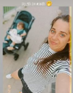 Sieviete svītrainā kreklā smaida pašbildē, bet fonā bērns sēž ratiņos; augšā redzams uzraksts un smaidiņš.