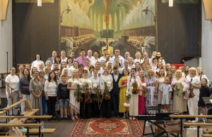 Kora un orķestra dalībnieki pēc koncerta pozē kopbildei baznīcā; priekšplānā mūziķes tur vijoles un ziedus.