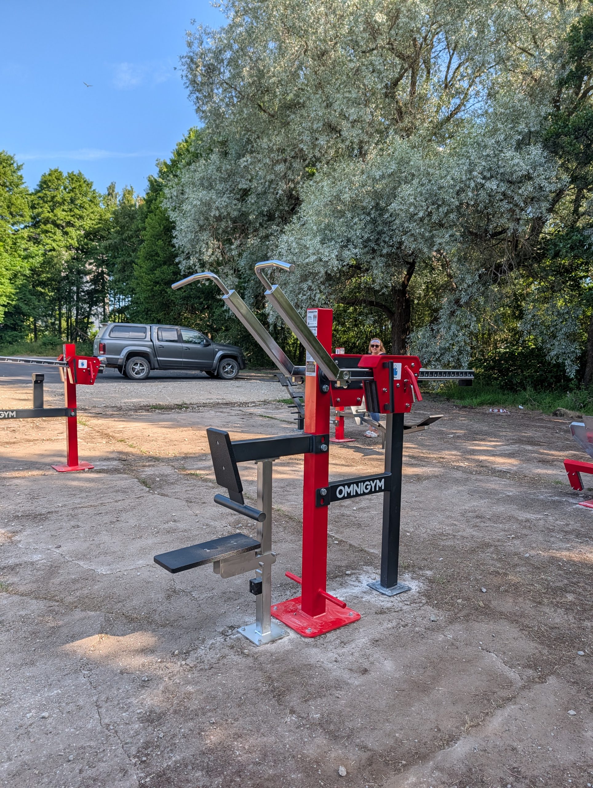 Āra trenažieru laukumā pie kokiem uzstādīti sarkani un melni spēka trenažieri; fonā redzama automašīna un cilvēks.