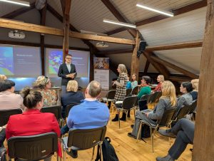 Semināra dalībniece uzrunā auditoriju koka siju zālē, kamēr vadītājs stāv pie ekrāniem ar prezentācijas slaidiem.