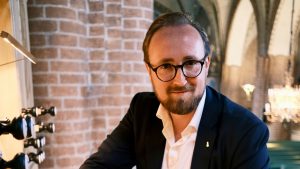 Ērģelnieka portrets pie ērģeļu reģistriem baznīcas interjerā ar ķieģeļu sienu un arkām fonā.