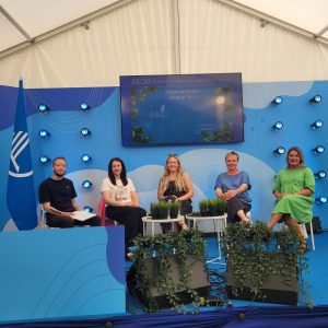 Pieci diskusijas dalībnieki sēž uz zilas festivāla skatuves ar augiem; ekrānā redzama tēma par transportu pēc pieprasījuma reģionos.
