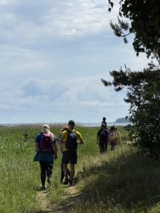 Pārgājiena dalībnieki ar mugursomām dodas pa šauru taku cauri piekrastes pļavai, fonā redzama jūra un mežains zemesrags.