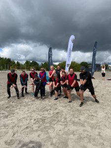 Ūdens soļošanas dalībnieki sarkanās vestēs pludmalē pozē vingrojuma pozā pie pasākuma karogiem zem tumšām debesīm.