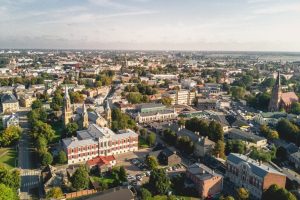 Jelgavas pilsētas panorāma no augšas ar baznīcu torņiem, vēsturiskām ēkām, zaļām koku alejām un plašu skatu uz pilsētu.