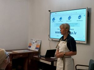 Semināra lektore priekšautā stāsta dalībniekiem pie ekrāna, kur redzama Baltijas ūdenstūrisma platforma “Upesoga”.