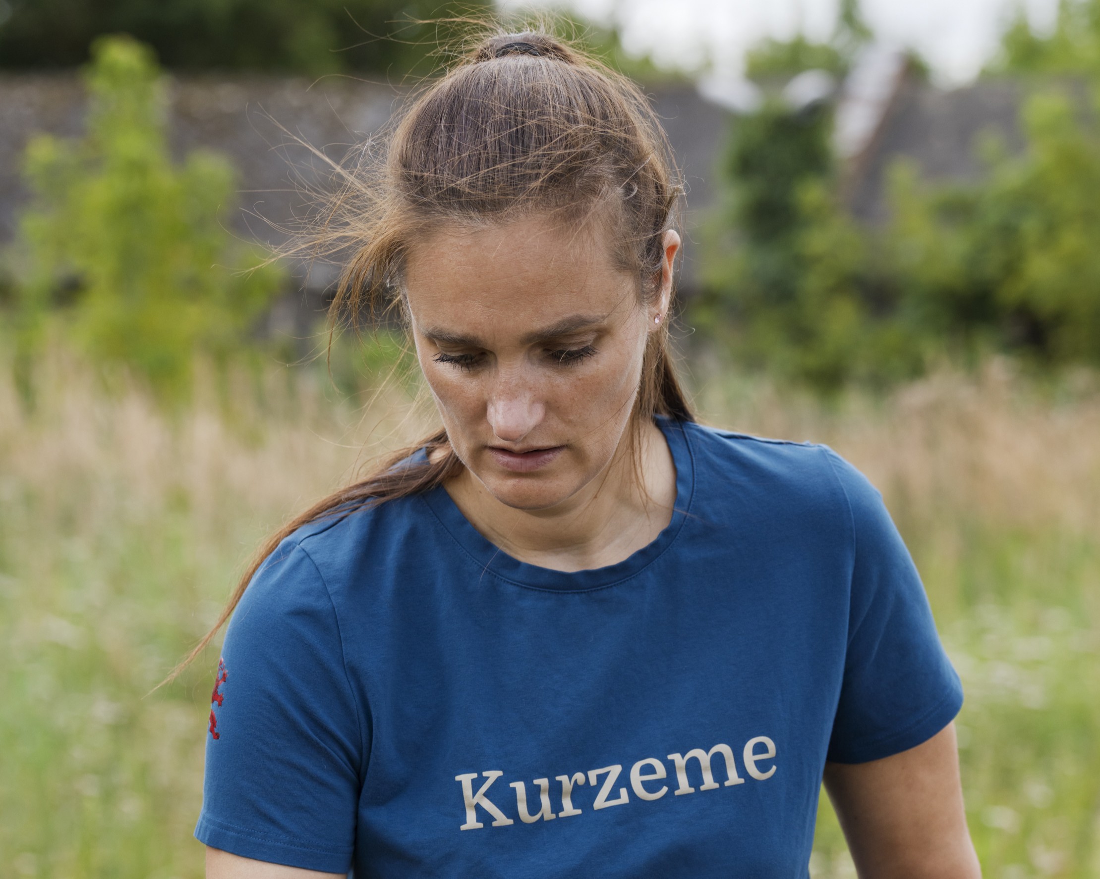 Sieviete zilā T kreklā ar uzrakstu “Kurzeme” stāv brīvā dabā un skatās lejup; fonā redzama izplūdusi zaļa ainava.