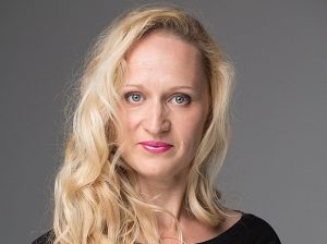Ērģelnieces Ilonas Birģeles studijas portrets: viņa ar gariem, viļņainiem blondiem matiem raugās kamerā uz pelēka fona.