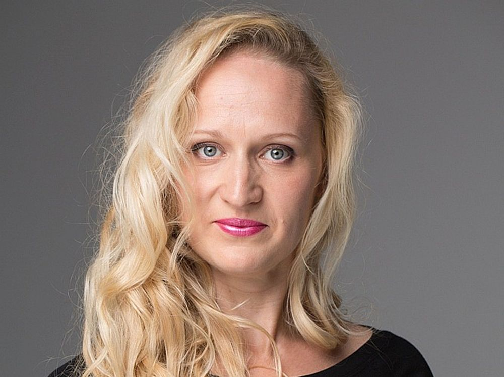Ērģelnieces Ilonas Birģeles portrets: gaišmataina sieviete melnā tērpā uz neitrāla pelēka fona skatās kamerā.