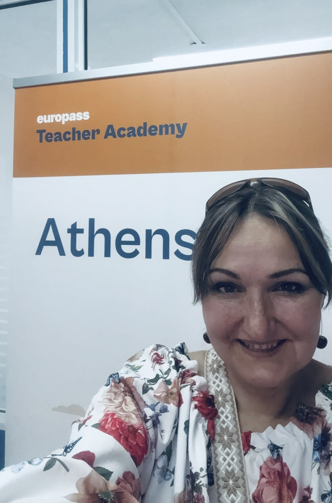 Mobilitātes dalībniece D. Kamela smaida pašportretā pie “Europass Teacher Academy” stenda Atēnās, tērpusies ziedu raksta blūzē.