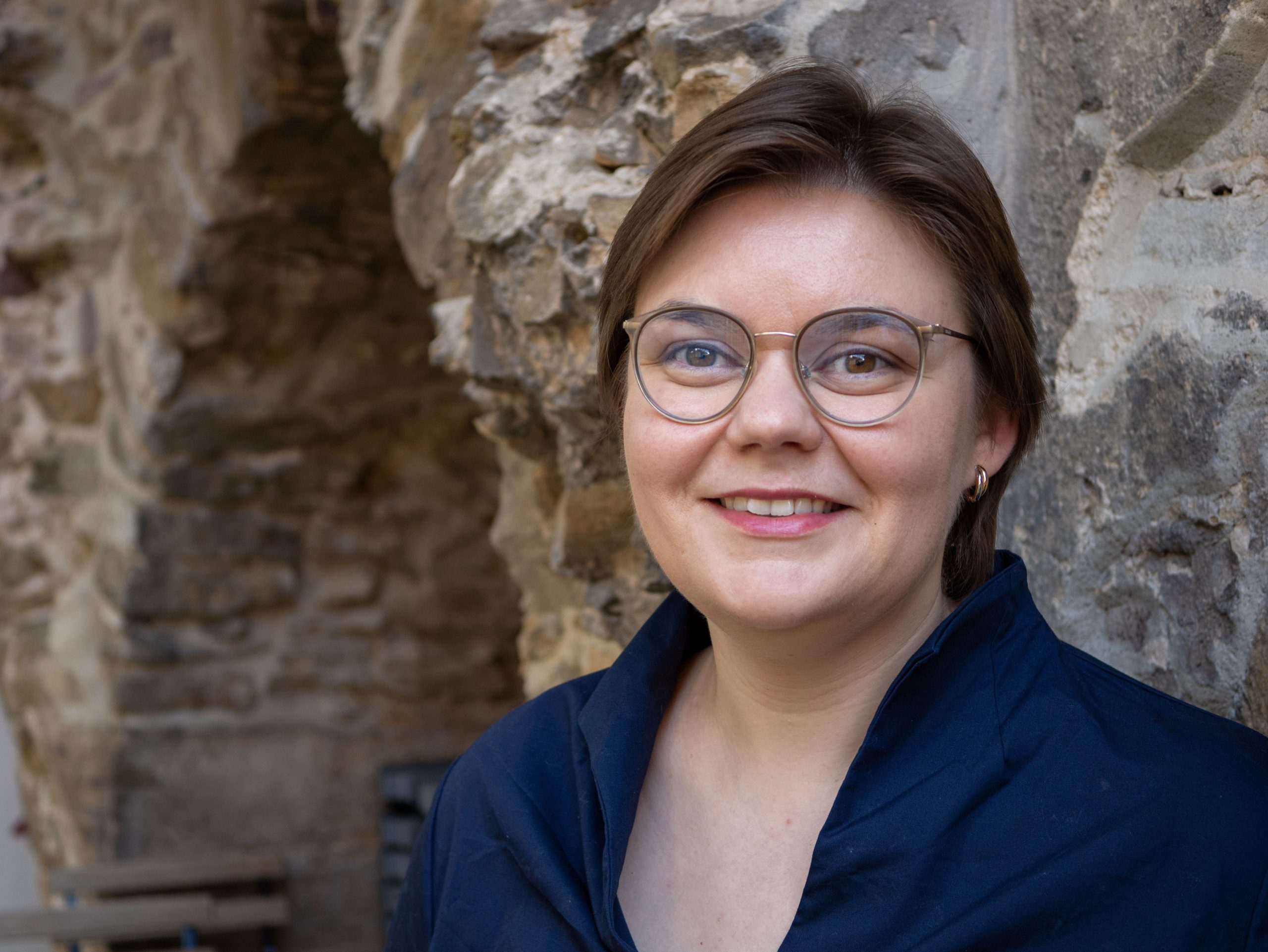 Ērģelniece Līga Auguste-Meijere tumši zilā tērpā un brillēs smaida pie senas akmens sienas, fonā redzama arka.
