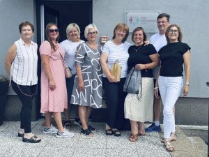 Astoņi mobilitātes dalībnieki smaidot pozē pie Čavles pensionāru kluba ieejas; viena dalībniece tur koka dēlīti.