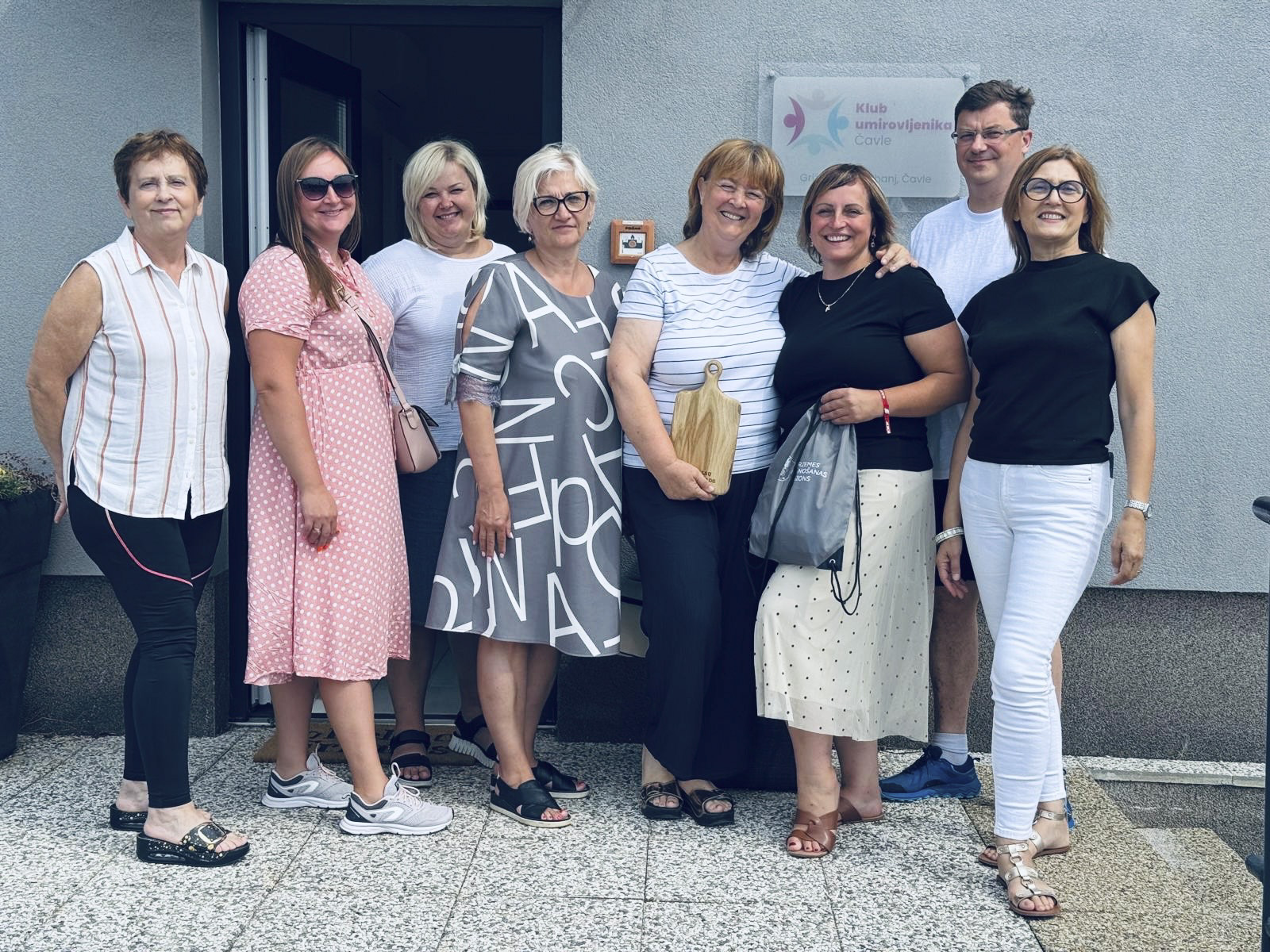 Astoņi mobilitātes dalībnieki smaidot pozē pie Čavles pensionāru kluba ieejas; viena dalībniece tur koka dēlīti.