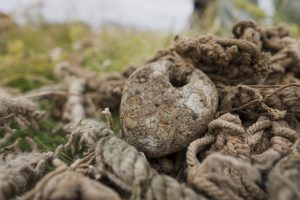 Tuvplānā starp vecām, sapinušām virvēm un zvejas tīkliem redzams apaļš, nodilis akmens vai smags tīklu atsvars ar caurumu.
