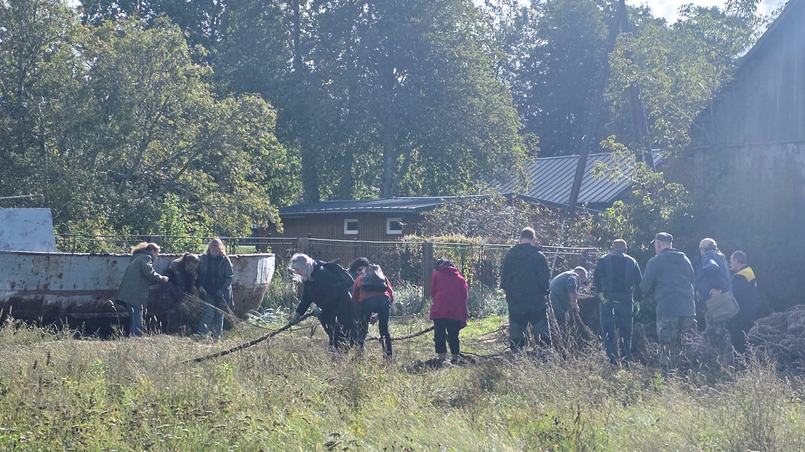 Cilvēku grupa strādā brīvdabas pļavā pie vecas laivas un ēkām, velkot virvi. Saulaina diena laukos ar kokiem fonā.