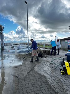 Cilvēki ostā tīra zvejas tīklus ar augstspiediena ūdens strūklu pie laivām.